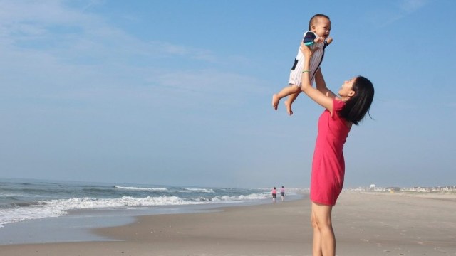 A mother holds her baby over head on a beach, which is one of the best places to have fun with babies.